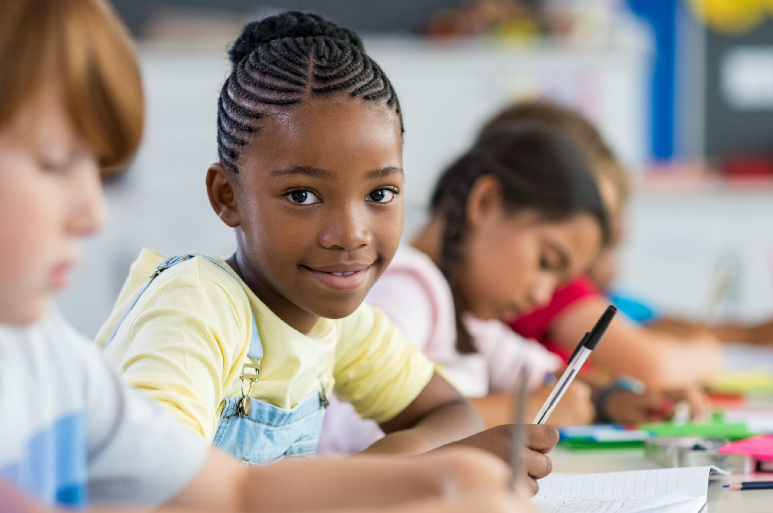 Smiling girl sitting at desk in class room and looking at camera. Portrait of young Black schoolgirl studying with classmates in background. Happy smiling pupil writing on notebook.