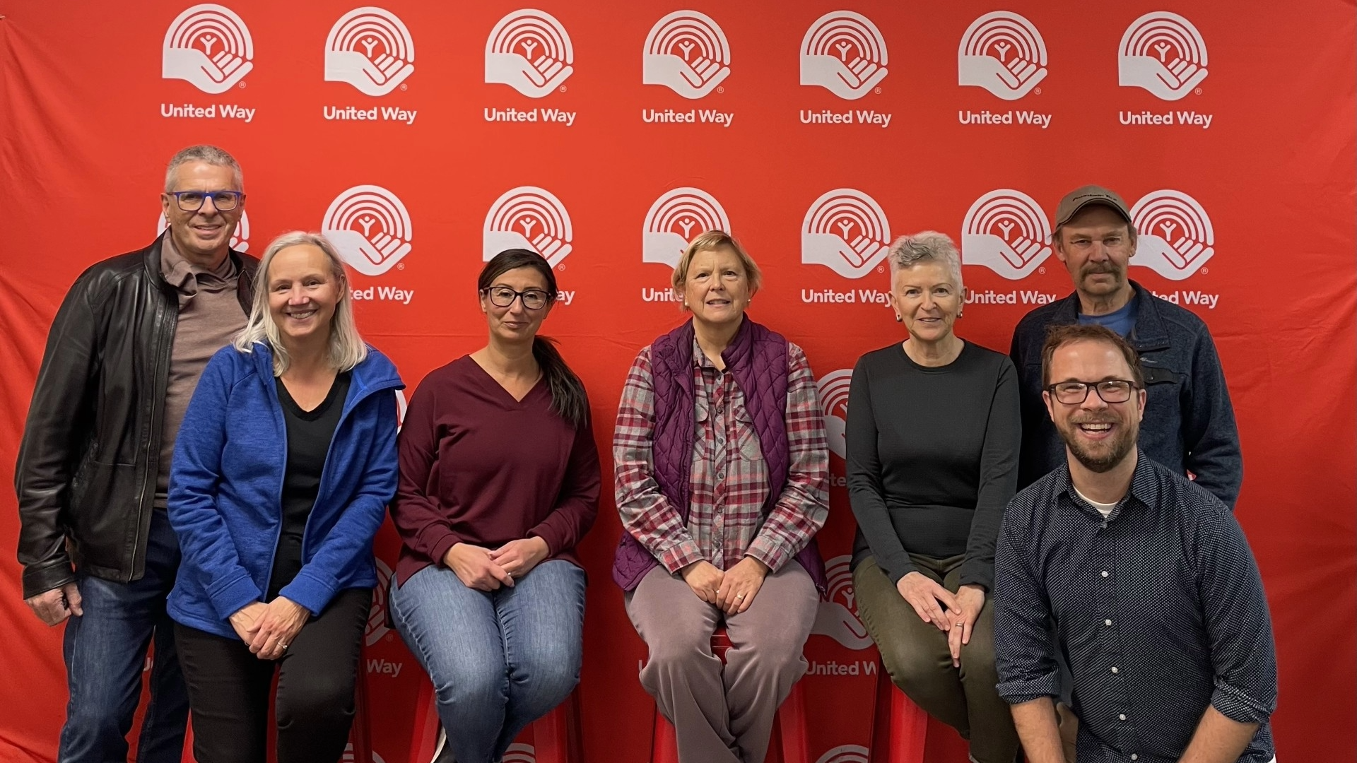 A group of ReUnited volunteers pose in front of a United Way backdrop. The group includes seven people of diverse ages, smiling proudly after participating in the Day of Caring event for the Coats for Kids & Families program.